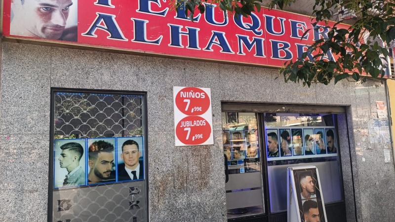 Fachada de Peluquería Alhambra: rótulo rojo con letras azules, escaparate con fotos de cortes y las pegatinas de precio para niños y jubilados.